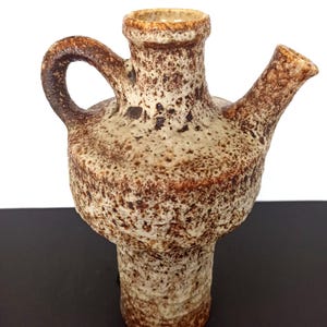 May include: A textured, ceramic pitcher with a handle and a spout. The pitcher is primarily beige with brown speckles and accents. The design features a wide base, a bulbous midsection, and a narrow neck. The handle is curved, and the spout extends horizontally.