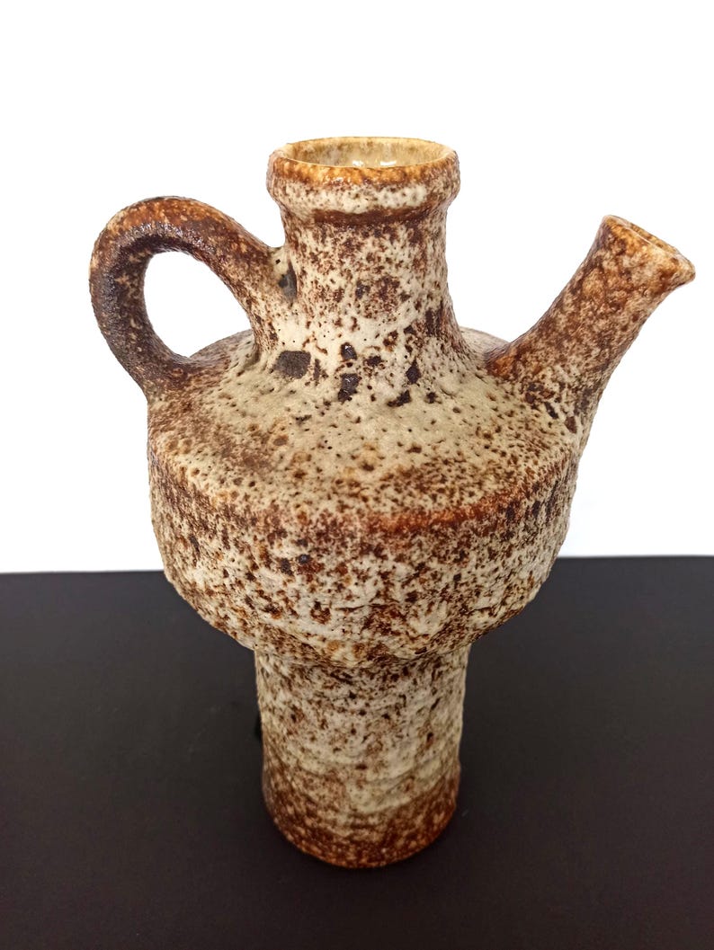May include: A textured, ceramic pitcher with a handle and a spout. The pitcher is primarily beige with brown speckles and accents. The design features a wide base, a bulbous midsection, and a narrow neck. The handle is curved, and the spout extends horizontally.