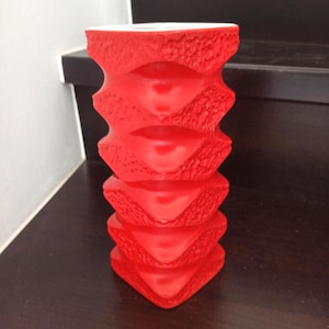 Könnte beinhalten: Eine leuchtend rote, strukturierte Vase mit einer einzigartigen Sanduhrform. Die Vase besteht aus einer Reihe von gestapelten, rautenförmigen Segmenten, die ein visuell auffälliges und modernes Design erzeugen. Das Innere ist weiß und kontrastiert mit der kräftigen Farbe des Äußeren.