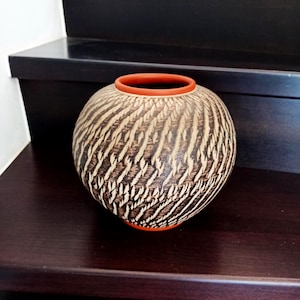 Könnte beinhalten: Eine kugelförmige Keramikvase mit einem terrakottafarbenen Rand und Boden. Die Außenfläche ist mit einem dunkelbraunen und cremefarbenen Texturmuster verziert, das aus sich wiederholenden diagonalen, geschnitzten oder geätzten federartigen Markierungen besteht.