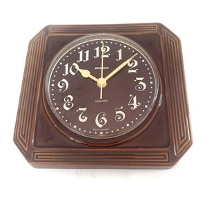 May include: A square brown ceramic wall clock with a glossy finish and stepped border. The dark brown face features white Arabic numerals, gold-tone hands, and the text "Stanger", "Quartz", and "West Germany" printed in white on the dial.