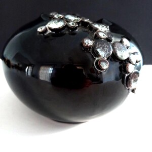 Könnte beinhalten: Eine glänzend schwarze Keramikvase mit abgerundeter, bauchiger Form. Die Oberfläche ist mit einer Ansammlung erhabener, kreisförmiger metallischer Akzente in Silber-, Bronze- und Dunkelgrautönen verziert, die einen strukturierten Kontrast zur glatten Oberfläche bilden.