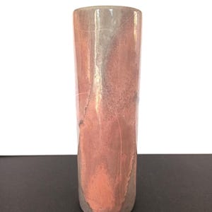 Könnte beinhalten: Eine zylindrische Vase mit strukturierter Oberfläche. Die Vase hat einen Farbverlauf aus Kupfer, Grau und rosa Farbtönen. Die Vase steht auf einer dunklen Oberfläche.
