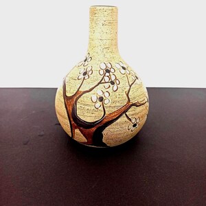May include: A beige ceramic vase with a round body and a narrow neck. The vase is decorated with a brown tree design and white flower blossoms. The vase is approximately 6 inches tall.