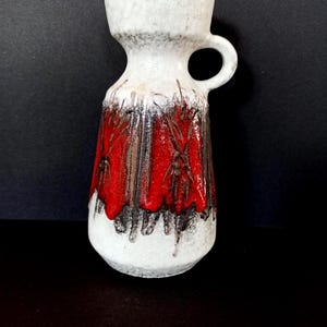 Puede incluir: Un jarrón de cerámica blanco y rojo con asa. El jarrón tiene una parte superior blanca texturizada y un diseño rojo y negro en la mitad inferior. El jarrón mide aproximadamente 18 cm de alto.