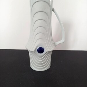 May include: A light grey ceramic mug with a distinctive, flared shape. The mug has a series of curved, wave-like ridges and a dark blue, circular accent. It has a handle and is suitable for beverages.