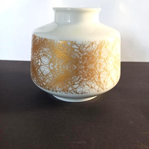 May include: A white ceramic vase with a gold, organic pattern. The vase has a rounded body and a narrow opening. The gold design covers the lower half of the vase, creating a striking contrast against the white background. The vase is likely for decorative purposes.