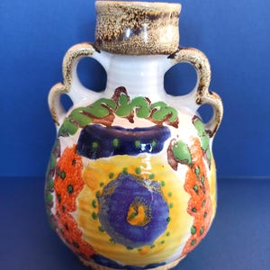 May include: A ceramic vase with two handles, featuring a white base decorated with hand-painted abstract patterns in yellow, orange, green, and dark blue. The rim and handles have a textured brown speckled finish, and the body is rounded.