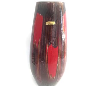 Puede incluir: Un jarrón de cerámica alto con un acabado brillante. El jarrón presenta una combinación de colores de rayas verticales rojas, marrones y marrón oscuro. Una pequeña pegatina dorada está adherida a la parte delantera del jarrón. El jarrón es un objeto decorativo.