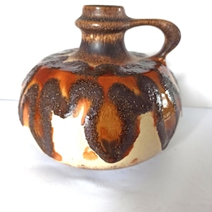 Puede incluir: Jarra de cerámica con cuerpo redondeado y asa. La jarra presenta un esmalte marrón y naranja con un diseño goteado sobre una base color crema. La jarra tiene un cuello corto y cilíndrico.