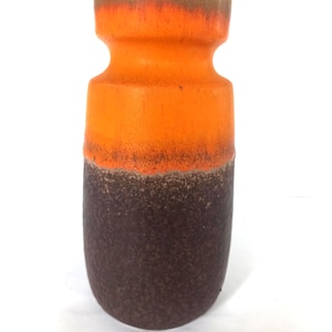 Può includere: Un vaso in ceramica bicolore. La parte superiore è arancione brillante, che sfuma in un marrone più scuro nella parte inferiore. Il vaso ha una superficie strutturata e una forma distintiva.