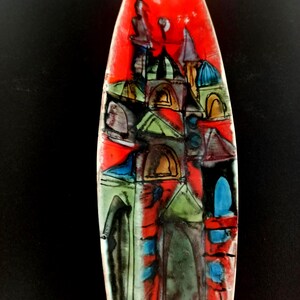 Puede incluir: Un jarrón de cerámica colorido con un diseño abstracto de paisaje urbano. La obra de arte presenta un fondo rojo con edificios verdes, azules y amarillos delineados en negro. El jarrón tiene una forma alargada.