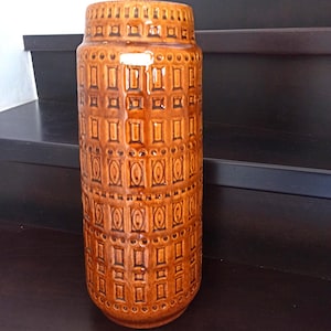 Op de afbeelding: Een hoge, cilindrische keramische vaas in een warme bruine tint. De vaas heeft een herhalend patroon van rechthoeken en cirkels, waardoor een textuur ontstaat. De boven- en onderranden hebben een decoratief ontwerp. Een decoratief stuk voor in huis.