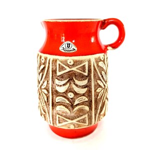 May include: A vintage ceramic jug with a bright red glaze on the top and base, and a textured, beige and brown patterned body. The jug has a handle and a small spout. The word "Keramik" is visible on a small sticker.