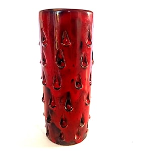 Può includere: Un vaso cilindrico in ceramica caratterizzato da una vivace smaltatura rossa con accenti scuri e maculati. La superficie è strutturata con numerose piccole rientranze a forma di goccia disposte in un motivo ripetuto sull'esterno.