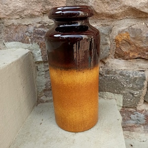 Pode incluir: Um vaso cilíndrico de cerâmica com um esmalte castanho escuro brilhante na parte superior e um acabamento texturizado amarelo-dourado mate na parte inferior. O vaso tem um rebordo largo e arredondado e um corpo reto e cilíndrico.