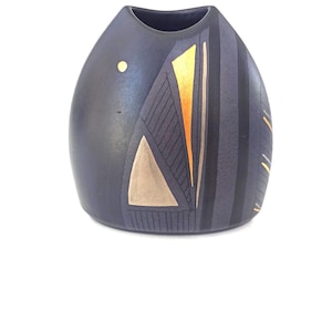 May include: A dark blue ceramic vase with a rounded shape and a narrow opening. The vase features gold and tan geometric designs on one side, including triangles and vertical stripes. A small gold dot is on the front.