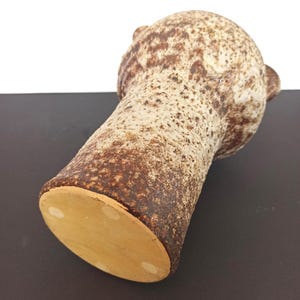 May include: A textured, brown and beige ceramic vase with a bulbous top and a cylindrical body. The vase has a speckled pattern and small, ear-like handles on the top. The base is a light tan color.