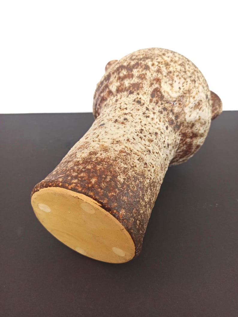 May include: A textured, brown and beige ceramic vase with a bulbous top and a cylindrical body. The vase has a speckled pattern and small, ear-like handles on the top. The base is a light tan color.