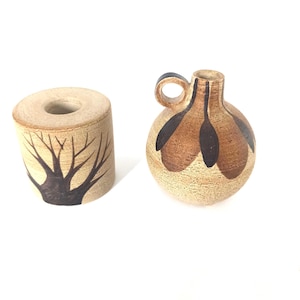May include: Two handmade ceramic pieces. One is a cylindrical candle holder with a tree branch design in dark brown. The other is a small, round pitcher with a handle and brown and black leaf-like patterns.
