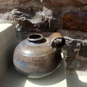 Könnte beinhalten: Ein handgefertigter, runder Keramikkanister mit dunkelbrauner und blauer Glasur. Der Krug hat eine kleine, kreisförmige Öffnung oben und einen gebogenen Griff. Die Keramik steht vor einer Steinmauer.