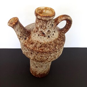 May include: A textured, brown and beige ceramic pitcher with a spout and handle. The pitcher has a bulbous body and a cylindrical base. The opening is a light beige color. The pitcher is likely vintage or handmade.