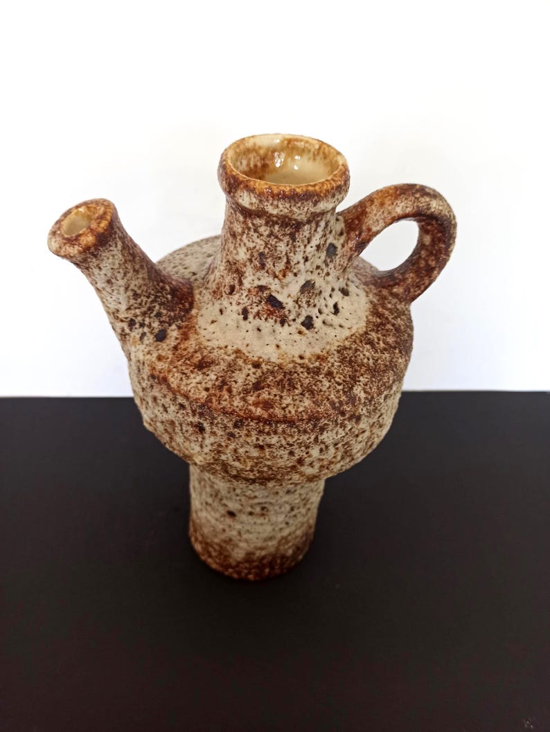 May include: A textured, brown and beige ceramic pitcher with a spout and handle. The pitcher has a bulbous body and a cylindrical base. The opening is a light beige color. The pitcher is likely vintage or handmade.