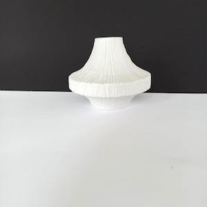 May include: A white ceramic vase with a textured, ribbed design. The vase has a wide, tiered base and a flared top. The background is a stark contrast of black and white, highlighting the vase's form and texture. This decorative piece is suitable for home decor.