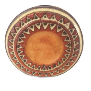 May include: Round ceramic dish with a central orange circle, surrounded by concentric rings. The rings feature a repeating triangle pattern in brown and cream against a reddish-orange background. The dish has a dark brown rim.