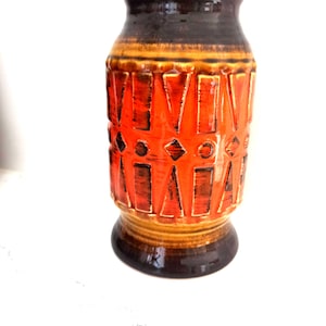 May include: A ceramic vase with a wide base and a narrow neck. The vase features a geometric pattern in shades of orange, brown, and yellow. The top and bottom are dark brown, with a lighter brown and yellow band in the middle.