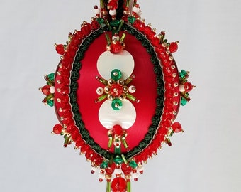 May include: A red and green beaded Christmas ornament with a white satin center and a white ribbon hanger.