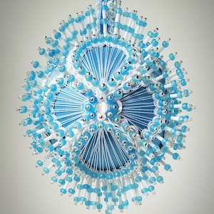 May include: A handcrafted, round ornament with a light blue fabric base, adorned with white thread, clear and light blue beads, and silver accents. The ornament is suspended by a light blue ribbon, creating a delicate, festive appearance.