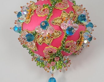 Könnte beinhalten: Ein rosa Weihnachtsbaumschmuck mit goldenen, grünen und blauen Verzierungen. Der Schmuck ist mit Perlen und Pailletten bedeckt und hat eine baumelnde blaue Perle.