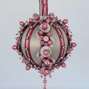 Puede incluir: Adorno navideño decorativo en tonos beige y burdeos. La bola redonda está adornada con cuentas rosas, detalles plateados y adornos florales. Una cinta burdeos está unida para colgarla, con un diámetro de unos 10 cm.
