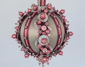 Könnte beinhalten: Ein dekorativer Weihnachtsschmuck in Beige und Burgunderrot. Der runde Schmuck ist mit rosa Perlen, silbernen Akzenten und floralen Verzierungen versehen. Ein burgunderfarbenes Band ist zum Aufhängen befestigt, mit einem Durchmesser von ca. 10 cm.