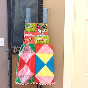 Può includere: Un grembiule in patchwork rosso, rosa, giallo, blu e verde con un'applicazione a forma di testa di volpe e le parole "New Forest" stampate su di esso.