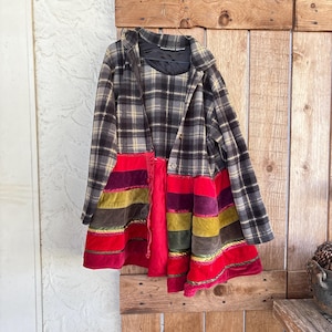 Op de afbeelding: Een unieke, handgemaakte jas met een geruit bovenstuk en een levendig, gestreept onderstuk. De jas heeft een mix van kleuren, waaronder rood, paars, geel en groen, en wordt getoond tegen een rustieke houten deur.