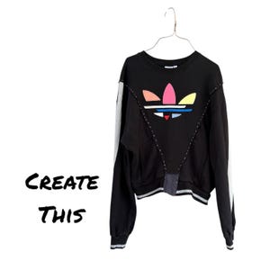 May include: Black sweatshirt with a colourful logo and white stripes on the sleeves. The text "CREATE THIS" is on the left side of the image. The sweatshirt is hanging on a metal hanger.