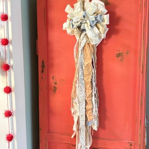 May include: A handmade fabric wall hanging with a cluster of cream and light blue fabric flowers at the top. Long, flowing ribbons in cream, tan, and lace cascade down. The wall hanging is attached to a distressed coral-colored cabinet.