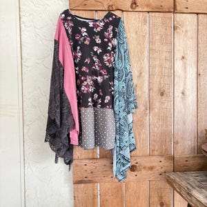 May include: A long-sleeved top with a patchwork design. The top features a black floral print, pink panel, gray polka dot section, and a blue paisley sleeve. A black lace panel hangs on the left side. The top is hanging on a wooden door.