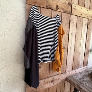Puede incluir: Una camisa de manga larga con un cuerpo de rayas horizontales blancas y negras, una manga gris oscuro lisa en el lado izquierdo y una manga amarilla mostaza con pequeños lunares negros en el lado derecho, colgada en una percha de alambre.