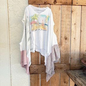 Puede incluir: Top blanco con cuello en V con un gráfico de escena de playa y el texto "Let's go to paradise". La parte superior tiene capas asimétricas con detalles en rosa y lunares negros. Cuelga de una puerta de madera.