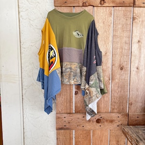 Puede incluir: Una camiseta patchwork con un diseño único. La camiseta presenta una combinación de colores y patrones, incluyendo verde oliva, camuflaje, azul y amarillo. Las mangas son de diferentes colores y patrones. La camiseta está colgada en una puerta de madera.