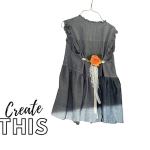 May include: A sleeveless, gray denim dress with a ruffled neckline and hem, hanging on a wire hanger. The dress features a gradient color effect, fading from gray to blue. A peach-colored rose and white lace accent the back. The text "Create THIS" is in the lower left.