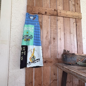 Op de afbeelding: Een patchwork tanktop gemaakt van hergebruikte t-shirts, met blauwe strepen, een vredesteken en het woord "HANG OUT". De tanktop hangt aan een metalen hanger tegen een houten deur.