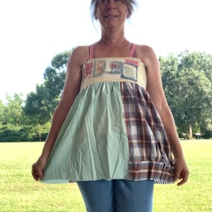 May include: A woman wearing a light blue and brown plaid top with a patchwork design. The top has a white band with a floral pattern and a pink and white striped band. The woman is wearing blue jeans.