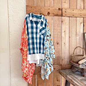 May include: A patchwork shirt with a blue and white plaid center, orange floral print on the left, and a light blue floral print on the right. The shirt has a white lace trim and is hanging on a wooden door.