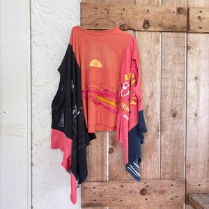 May include: A patchwork top constructed from multiple t-shirts in coral, black, and red. The front features a graphic of a sun over waves with layered fabric strips. The garment includes text elements like Cozumel and Rip Curl, with an asymmetrical hem.