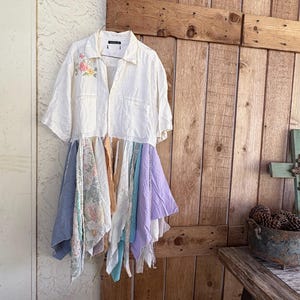 Può includere: Un abito camicia bianco a maniche corte, con ricami floreali e una gonna patchwork di vari tessuti pastello. L'abito è appeso a una gruccia contro una porta di legno rustica.