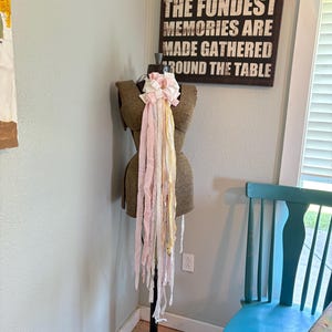 May include: A brown dress form with a decorative bow and fabric streamers in shades of pink, white, and yellow. The dress form is on a black metal stand. A sign on the wall reads "THE FONDEST MEMORIES ARE MADE GATHERED AROUND THE TABLE."